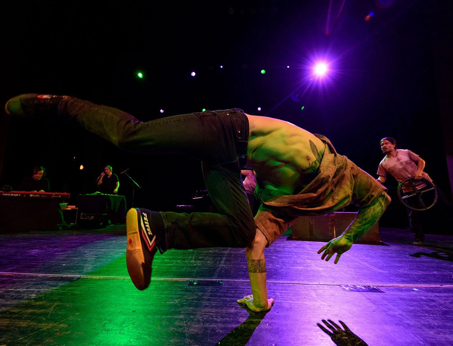 Ein Breakdancer balanciert auf einer Hand, die Beine seitlich ausgestreckt. Hinter ihm spielen Musiker im grünen und violetten Bühnenlicht.