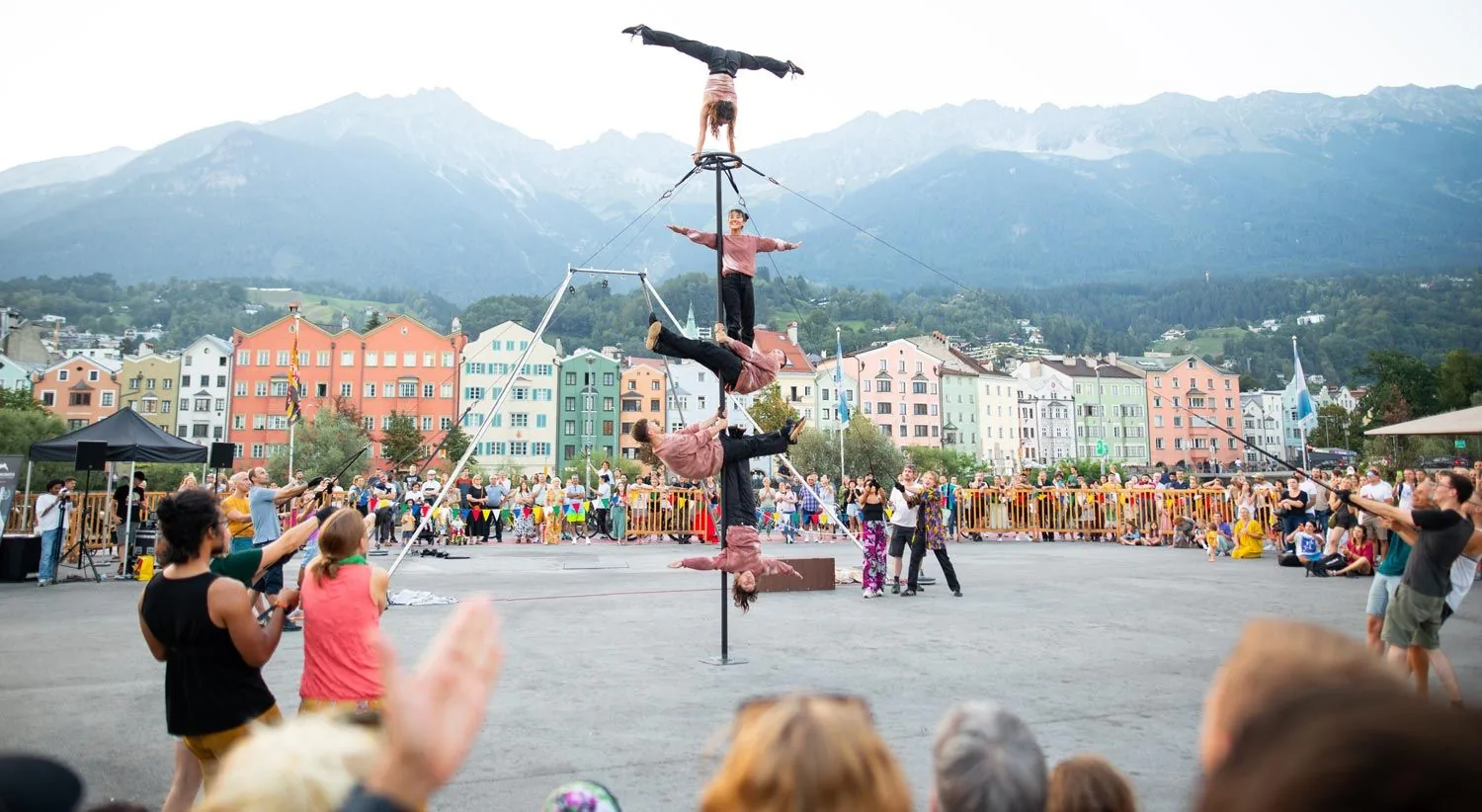 Mehrere Akrobaten halten verschiedene Positionen übereinander an einem Mast. Rundherum steht Publikum, dahinter liegen bunte Häuser und die Berge von Innsbruck.